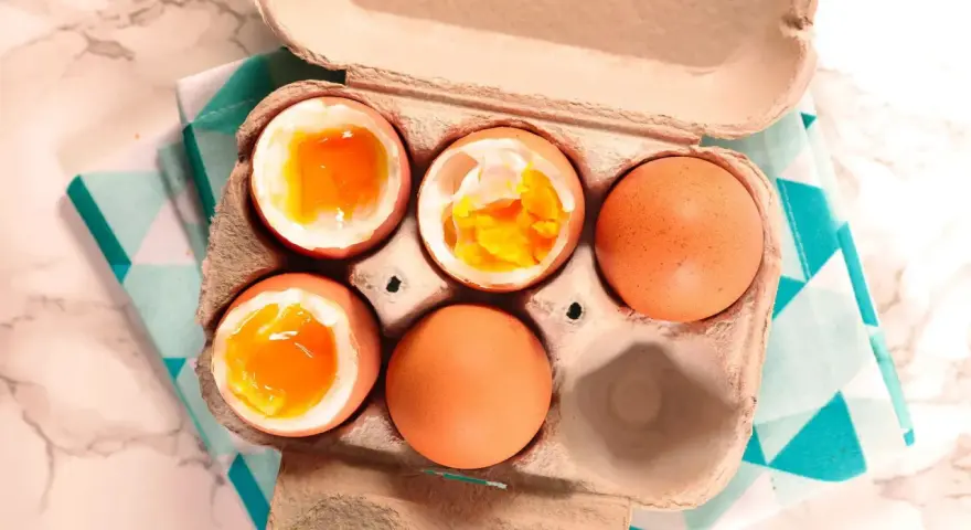 Uova alla coque in un cartone, alcune aperte mostrano tuorli dorati e cremosi, pronte per essere gustate.