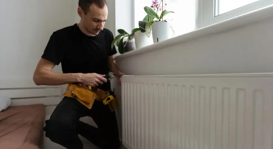 A man in a tool belt inspects a white radiator, checking for any signs of a cracked radiator before winter sets in.