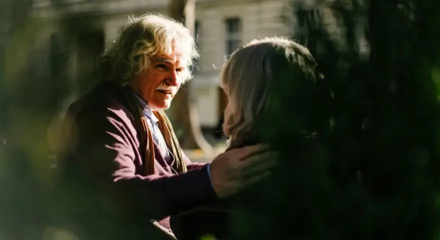 A man with white, curly hair and a mustache gently places his hand on another person's shoulder, offering a comforting response. This scene illustrates react vs respond examples.