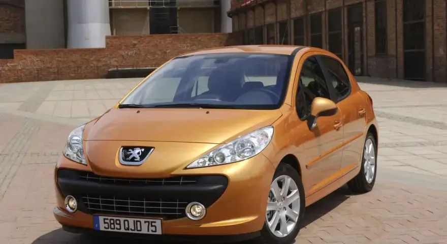 Pomarańczowy Peugeot 207 1.4 benzyna zaparkowany na brukowanym placu przed ceglanym budynkiem.