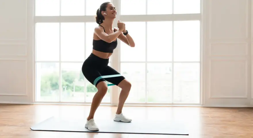 Femme souriante faisant un squat avec un élastique autour des cuisses, sur un tapis de yoga, devant une grande fenêtre lumineuse.