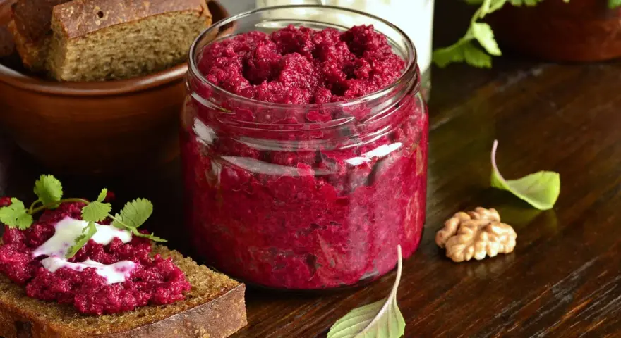 Glas mit leuchtend rotem Rote Bete Pesto, daneben Brot mit Pesto und Joghurt. Walnuss und Kräuter garnieren die Szene.