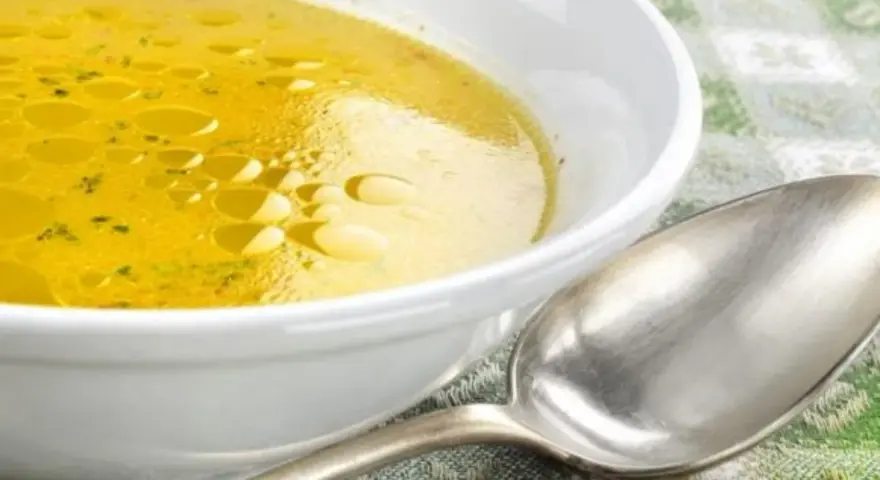 Una ciotola bianca piena di brodo di carne dorato, con goccioline d'olio in superficie. Un cucchiaio d'argento è appoggiato accanto.