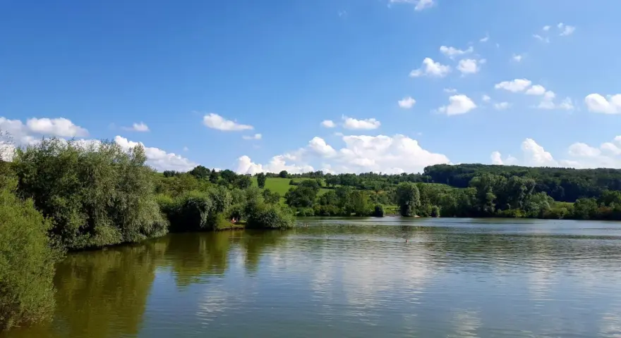 Ein sonniger Tag am Badesee Esslingen. Grüne Hügel, Bäume und ein blauer Himmel mit weißen Wolken spiegeln sich im ruhigen Wasser.