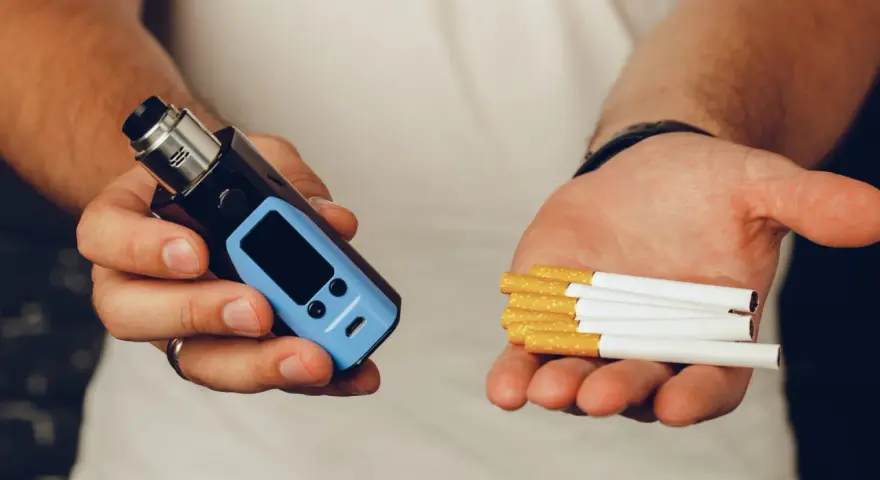 Man holds a vape in one hand and cigarettes in the other, posing the question: que es mejor el vaper o el tabaco.