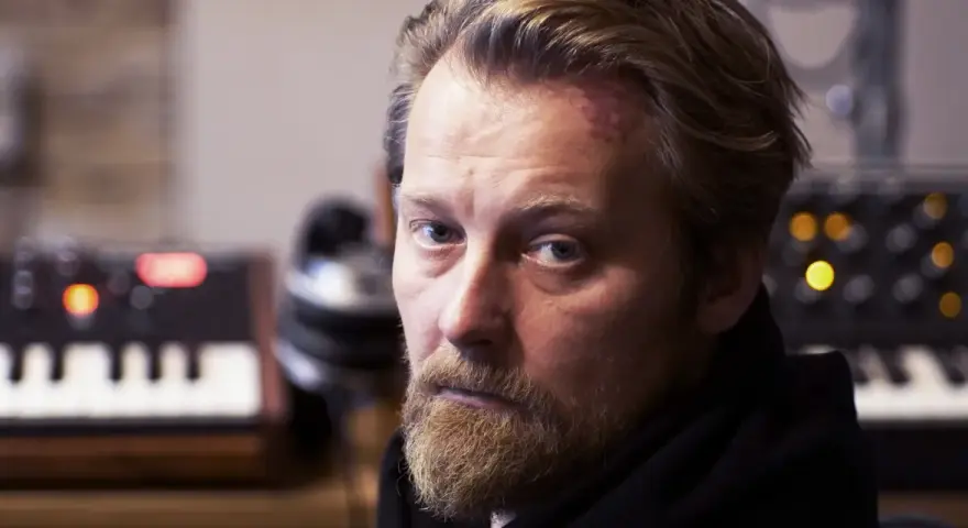 Bartek Dziedzic, z zarostem i lekko zaczerwienionym czołem, siedzi przed syntezatorami.