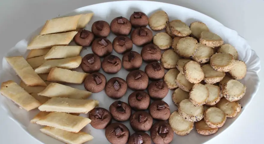 En vit oval bricka fylld med tre sorters hembakade småkakor: rombiska, chokladpraliner och runda med strössel. Perfekt för fika!