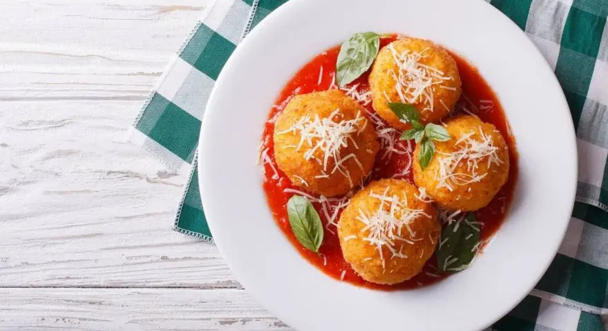 Quattro arancini di riso dorati, cosparsi di parmigiano e serviti su un letto di salsa di pomodoro, guarniti con basilico fresco.