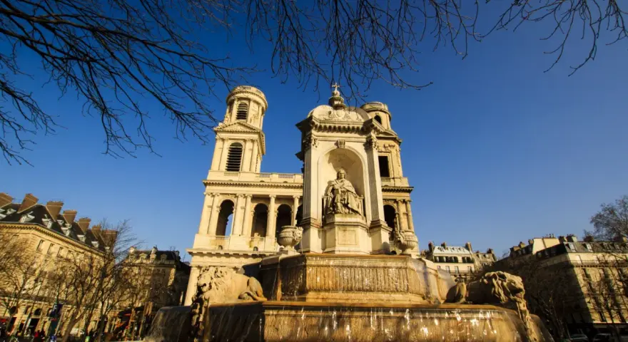 Fuente monumental frente a la iglesia de Saint Sulpice, con leones de piedra y una estatua central bajo un cielo azul claro.