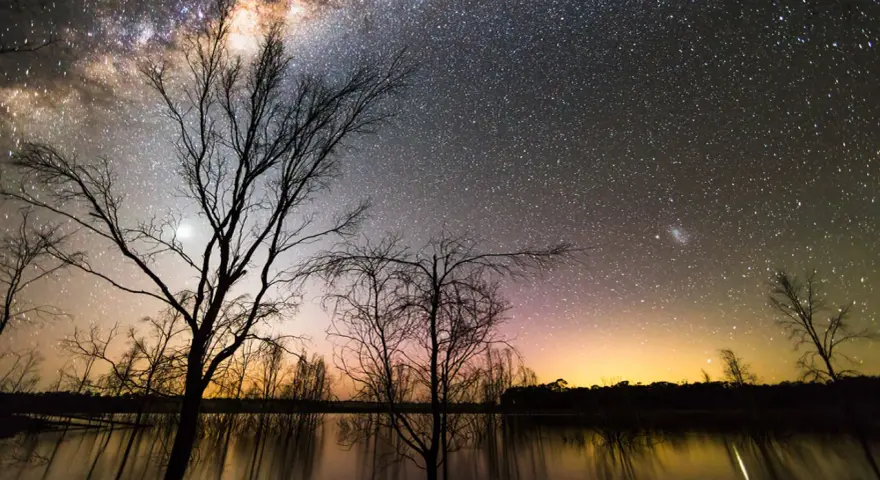Canon EOS 2000D photo de nuit : une voie lactée spectaculaire au-dessus d'un lac reflétant les arbres dénudés et un ciel orangé.