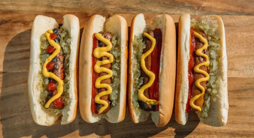Cuatro hot dogs con mostaza, kétchup y cebolla picada sobre una tabla de madera.