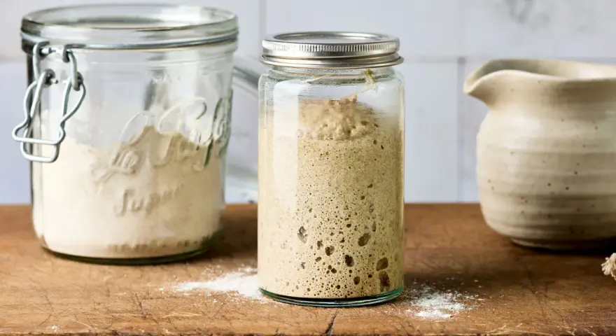 Blubbernder Sauerteig im Glas, bereit zum Backen. Daneben ein Glas mit Mehl, das zeigt, wie man den Sauerteig pflegen kann.