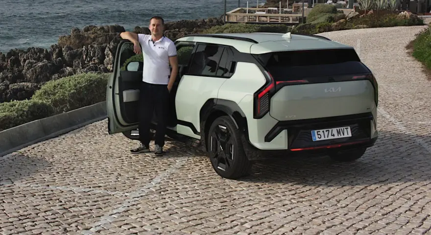 Mężczyzna opiera się o drzwi nowego, jasnoszarego Kia EV3, zaparkowanego na brukowanej drodze nad morzem.