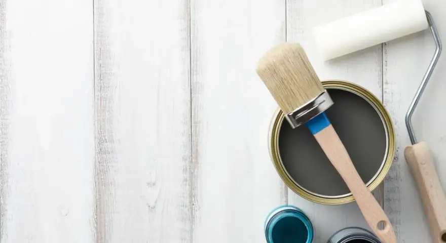 Herramientas para pintar mesitas de noche: lata de pintura gris, pincel, rodillo y botes de pintura azul y negra sobre fondo de madera blanca.