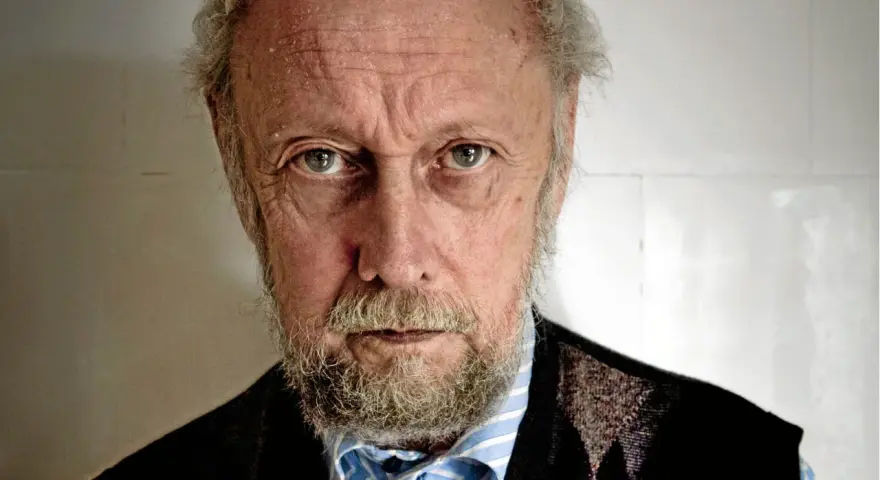 En äldre man med grått skägg och rynkor i ansiktet, som om han funderar på en handkraft novell.