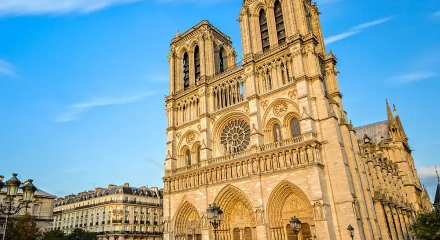 La majestueuse façade de Notre Dame de Paris se dresse sous un ciel bleu, ornée de détails gothiques. Des passants flânent sur la place.