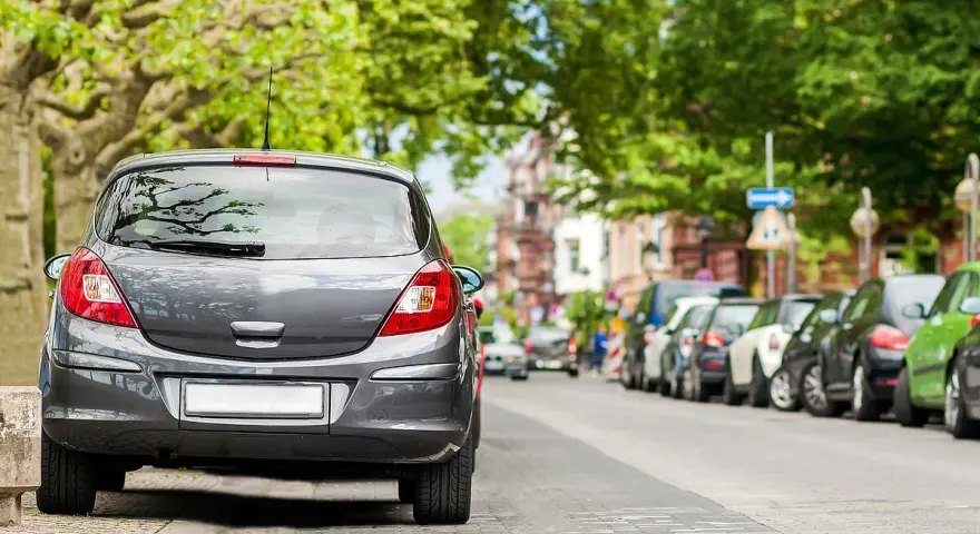 Graues Auto parkt am abgesenkten Bordstein. Mehrere Autos reihen sich die Straße entlang unter grünen Bäumen.