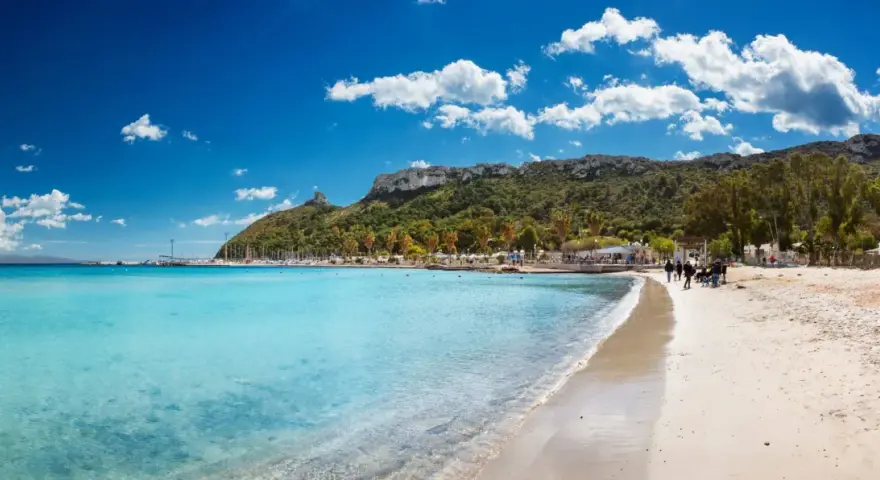 Spiaggia con acque cristalline e la sagoma della Sella del Diavolo all'orizzonte, sotto un cielo azzurro con nuvole bianche.