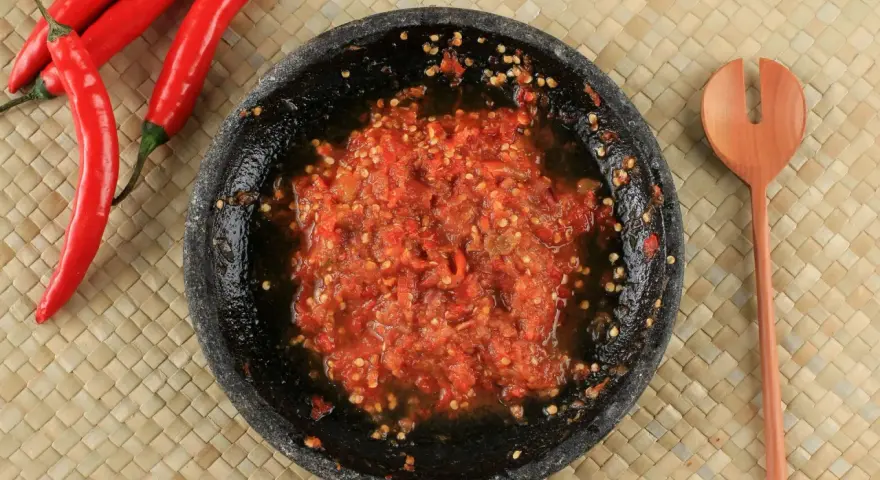 Świeżo przygotowany sos sambal w kamiennym moździerzu, obok czerwone papryczki chili i drewniana łyżka.