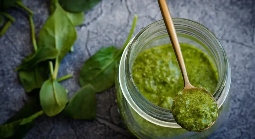 Słoik z zielonym pesto, na łyżce porcja sosu. Czy pesto ze słoika się podgrzewa? Świeże liście bazylii obok.