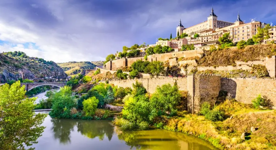 El **río Tajo** serpentea al pie de Toledo, una ciudad amurallada coronada por un imponente alcázar. Un puente de piedra cruza el agua, conectando las verdes orillas.
