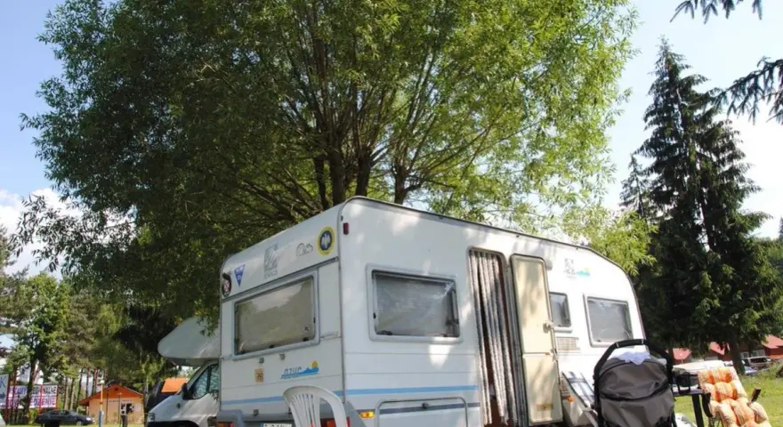 Przyczepa kempingowa na polu namiotowym Jonidło, otoczona zielenią. Obok wózek i leżak.
