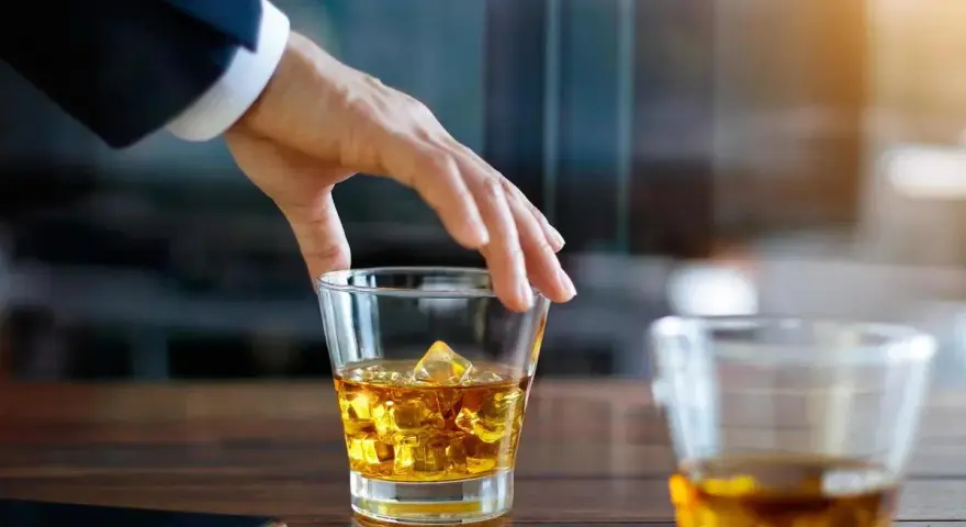 Hand greift nach einem Glas goldbraunen Whiskey mit Eiswürfeln. Ein Genuss, auch für Menschen, die glutenfreien Whiskey bevorzugen.