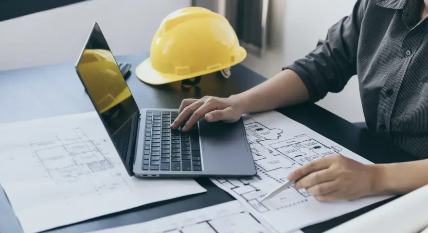 Architekt pracuje nad projektem, analizując plany i obliczając powierzchnię całkowitą budynku. Żółty kask ochronny leży obok.