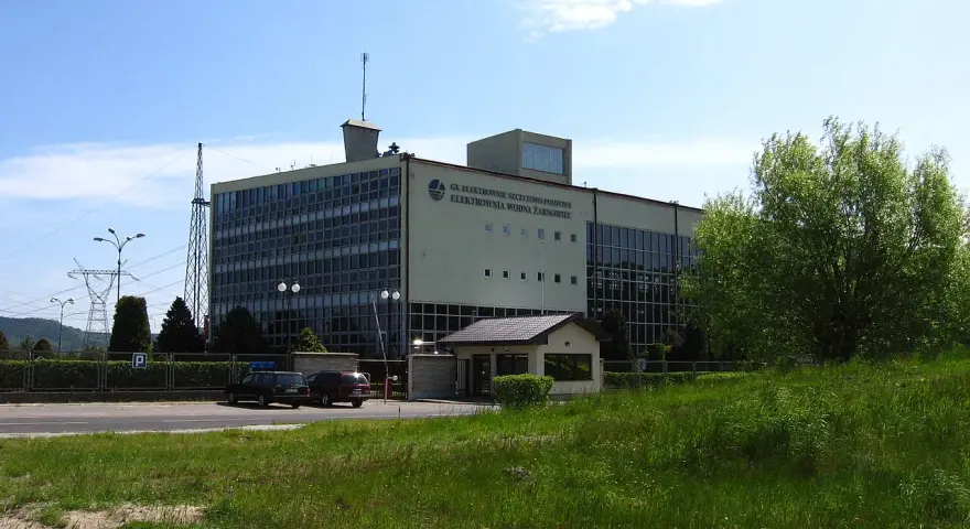 Budynek elektrowni wodnej Żarnowiec z charakterystycznym logo. W tle wieża transmisyjna i zieleń.