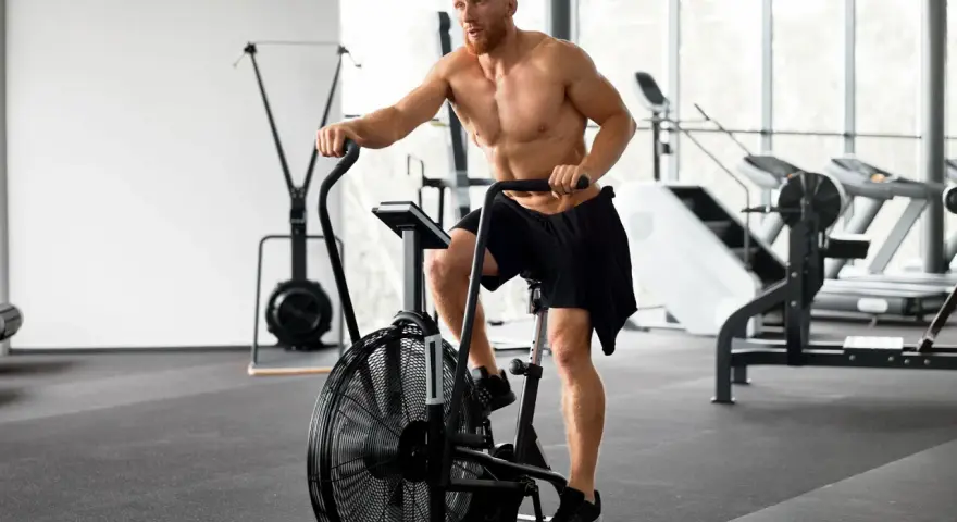 Mężczyzna ćwiczy na rowerze stacjonarnym typu air bike w siłowni.