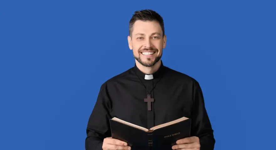 Uśmiechnięty ksiądz w czarnej sutannie z krzyżykiem trzyma otwartą Biblię. Idealny obrazek na podziękowania dla księdza.