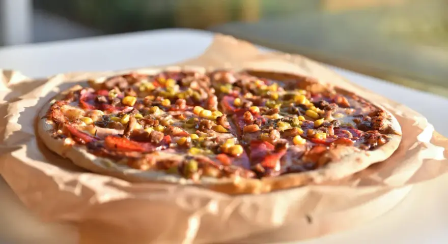 Apetyczna pizza Giuseppe 4 sery, z kukurydzą i mięsem, na papierze do pieczenia, w słonecznym świetle.