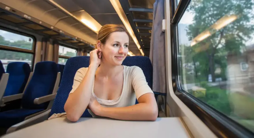 Uśmiechnięta kobieta w pociągu, patrząca przez okno. Czeka na kolejne przesiadki, ciesząc się podróżą.