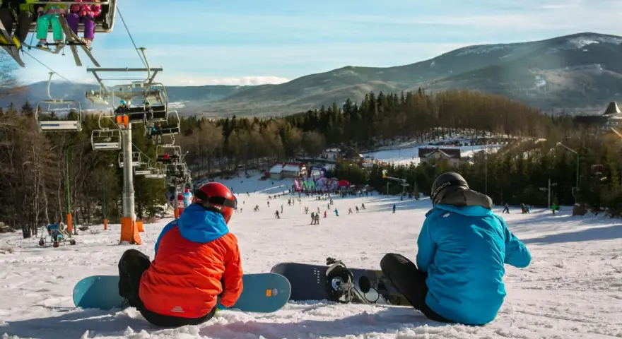 Dwaj snowboardziści odpoczywają na stoku, w tle widać wyciąg narciarski Karpacz i góry.