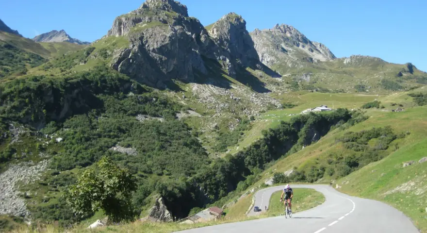 Ciclista su strada tortuosa verso il Cormet de Roselend, con montagne imponenti e verdi pendii.