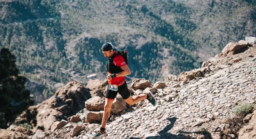 Un corredor de trail desciende por un sendero rocoso y empinado, con montañas boscosas al fondo.