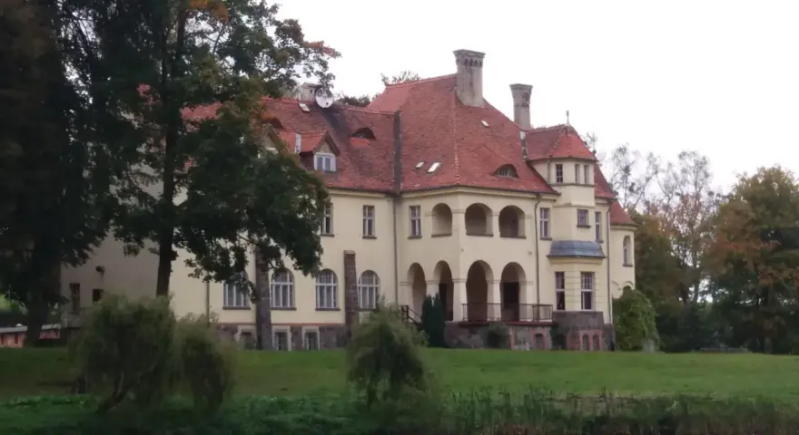 Malowniczy pałac z czerwoną, dachówkową, spadzistą dachem, otoczony zielenią i wodą.