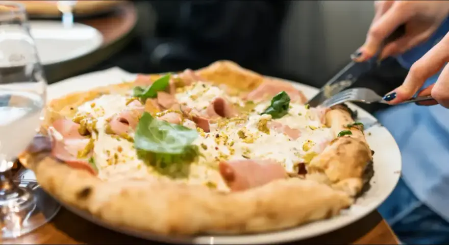 En person skär i en pizza bianco med skinka och basilika. Ett glas vatten står bredvid. Perfekt vin till pizza bianco.