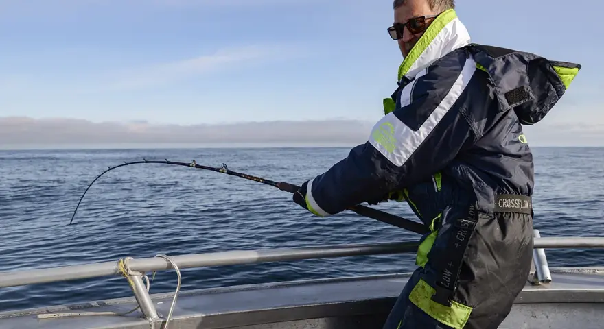 Man med solglasögon och fiskeutrustning kämpar med en böjd spö ombord på en båt. En spännande dag med fiska i Norge.