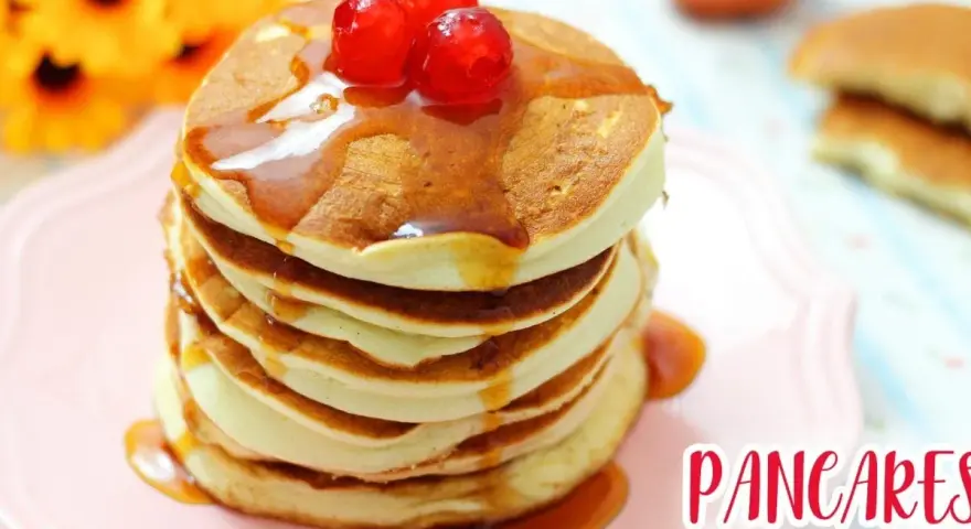 Pancakes senza glutine, soffici e dorati, impilati su un piatto rosa, con sciroppo d'acero e ciliegie rosse.