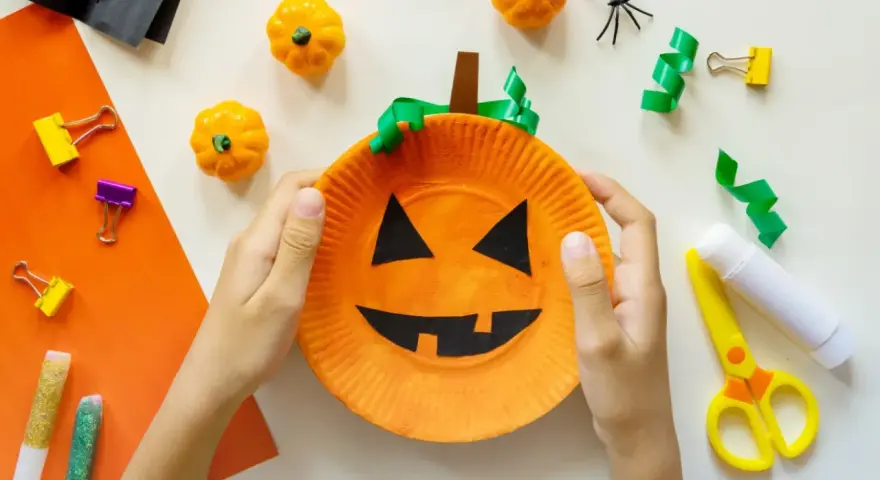 Barn gör halloween pyssel med en pumpa av papper. Små pumpor, spindlar och glitterpennor ligger redo.