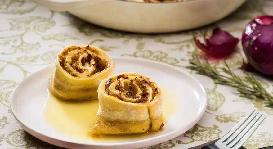 Zwei goldbraune Krautkrapfen im Sud auf einem Teller, im Hintergrund ein Auflauf mit weiteren Krapfen, Zwiebeln und Rosmarin. Ein leckeres Krautkrapfen Rezept.