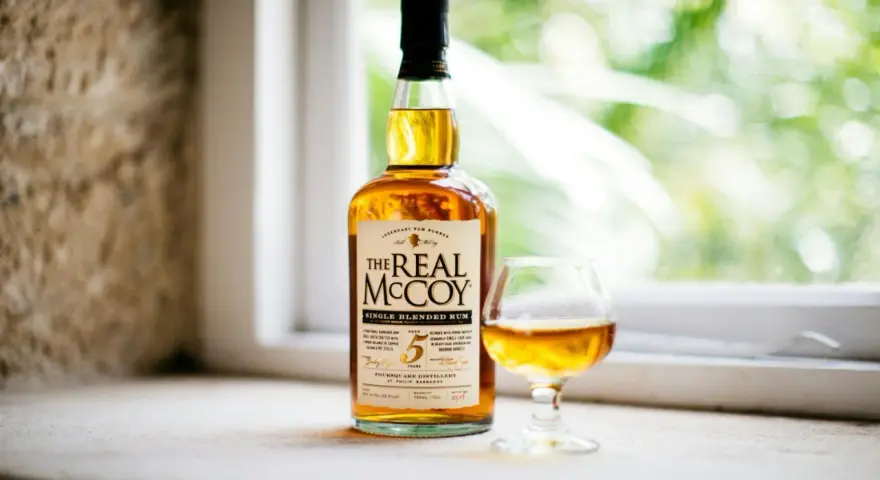 Ein besonderer Rum, "The Real McCoy", steht neben einem Glas mit bernsteinfarbener Flüssigkeit vor einem Fenster mit grünem Bokeh.