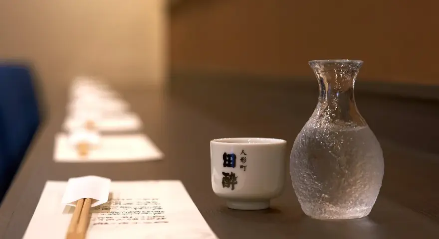 Sake ile procent? Delikatna czarka i karafka z sake czekają na degustację. W tle rząd nakryć.