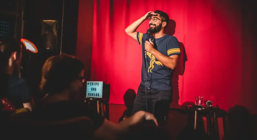 Ein Comedian steht auf einer Bühne in Berlin, hält ein Mikrofon und wischt sich die Stirn. Ein Schild mit "Chips und Kaviar" steht daneben.