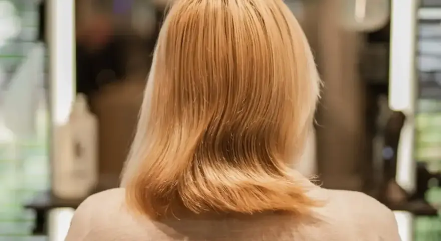 Blond włosy po zabiegu farba demi permanentna, delikatnie pofalowane, w tle rozmyte światła salonu fryzjerskiego.