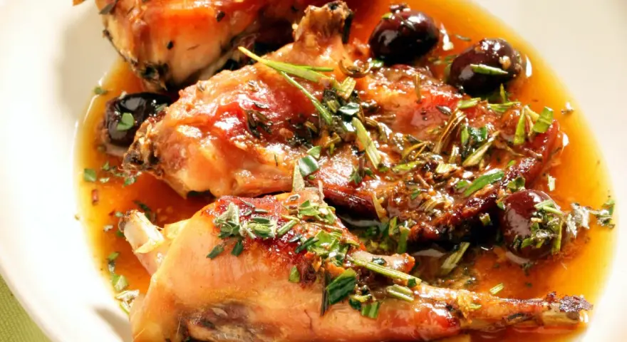 Coniglio alla ligure succulento, con olive nere e erbe aromatiche, servito in un piatto bianco.