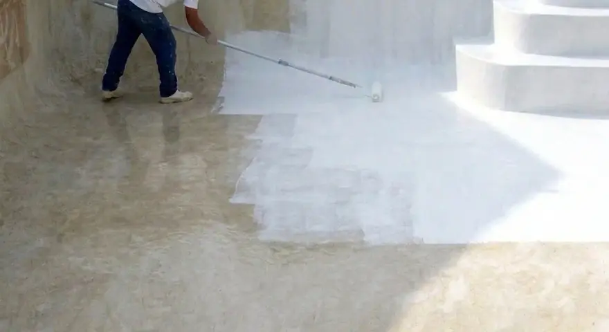 Hombre pintando una piscina de cemento con rodillo. La mitad de la piscina ya está pintada de blanco.