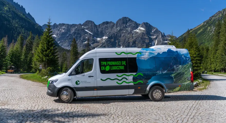 Biały bus Tatrzańskiego Parku Narodowego z grafiką górskiego krajobrazu, przypominającego Morskie Oko, stoi na kamiennej drodze.