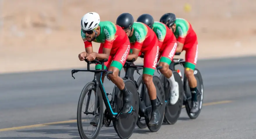 Équipe cycliste marocaine en tenue rouge et verte, concentrée sur la course. Ils roulent en peloton, tels des pur sang arabes noirs, sur un parcours désertique.